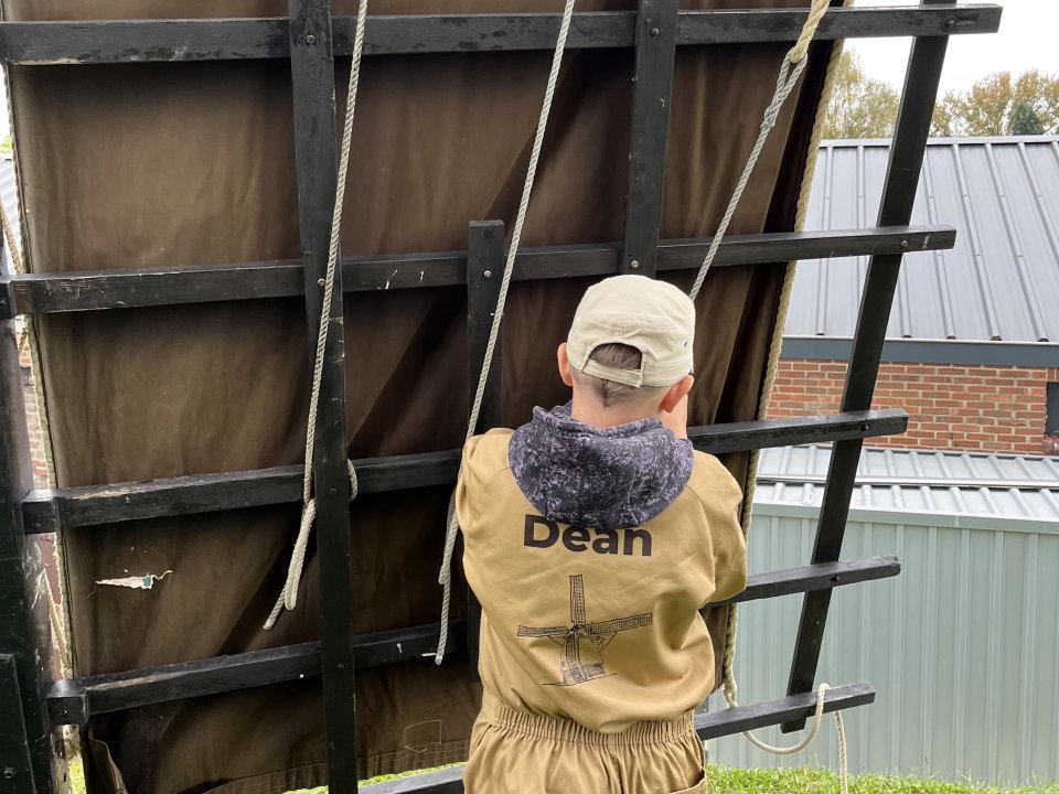 Rug van een jongen die bezig is de wiek van een molen op te zeilen. De jongen heeft een petje op en een beige overall aan met daarop de naam Dean