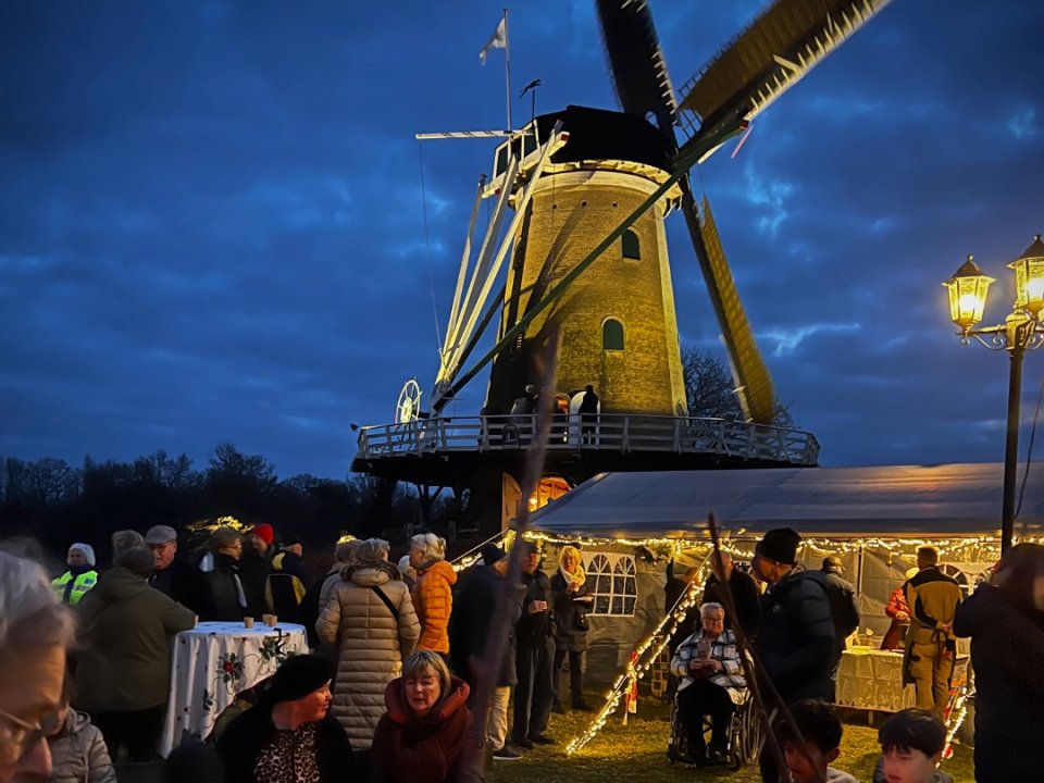 Avonmdfoto van kerstmarkt bij korenmolen De Windhond. Sfeervolle foto met veel mensen, een midwinterhoornblazer en marktkramen. 