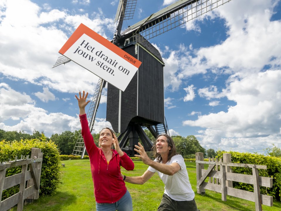 Staand beeld van molen in polderlandscha[p. op de voorgrond staan een man en een vrouw die een cheque vangen in de wind