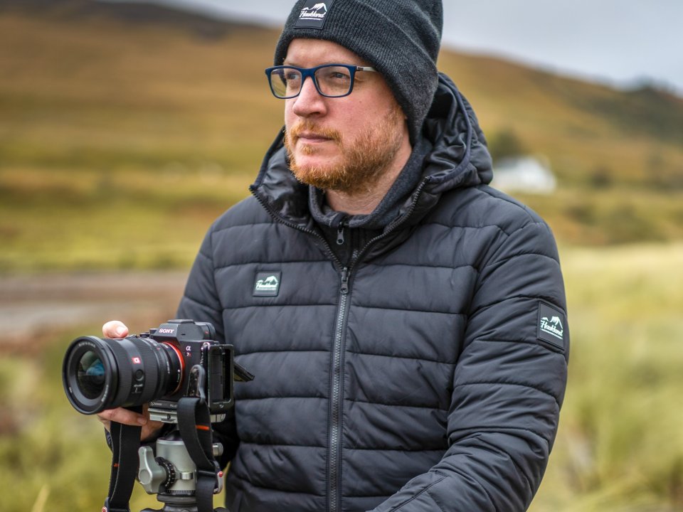 Portfretfoto van man met bril en zwarte muts op met spiegelreflexcamera in groen weids landschap