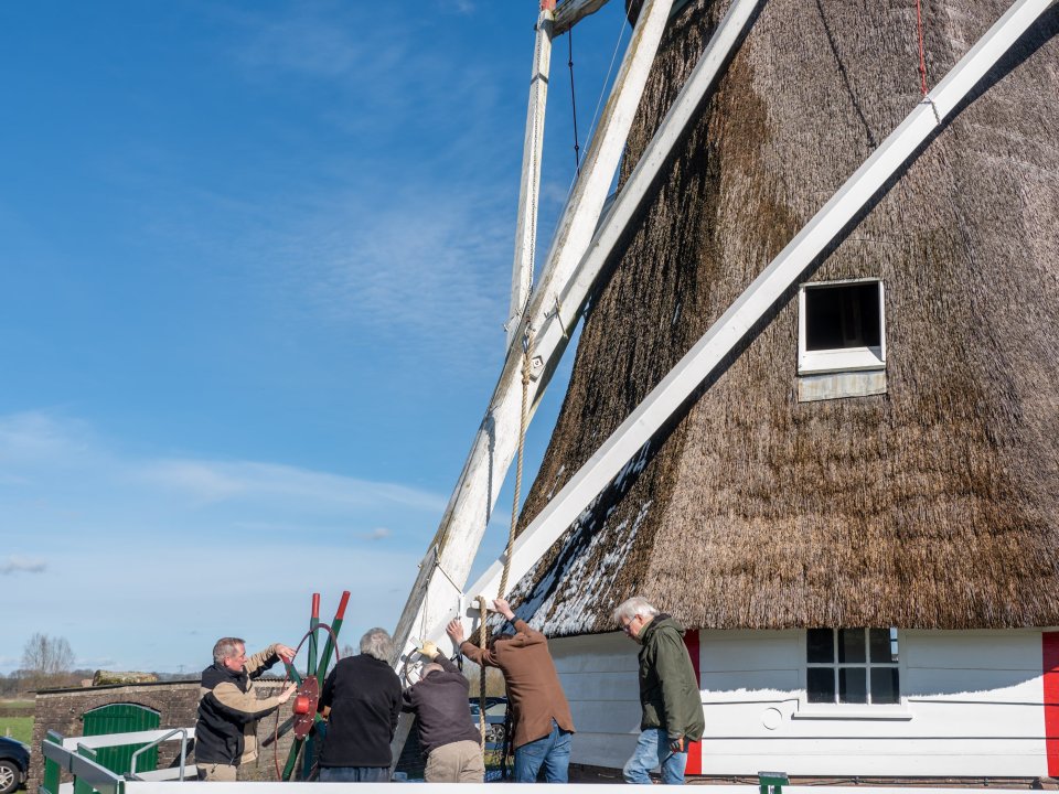 Deel van de Warkense Molen, bij de molen staat een groep van vier mannen bij het rad om proberen de molen te draaien (kruien).